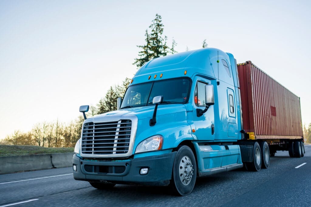 Front view of blue semi truck hauling commercial cargo container on highway – long haul transportation with green roadside scener